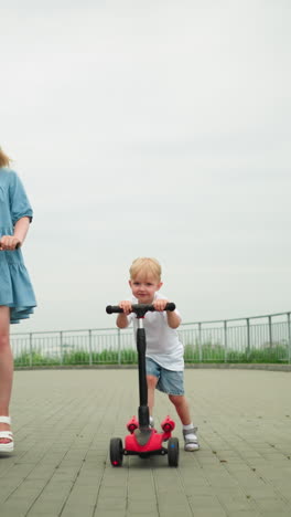 una madre y sus dos hijos montan scooters juntos en un área de parque abierto, con un carril de hierro al lado y bancos alrededor