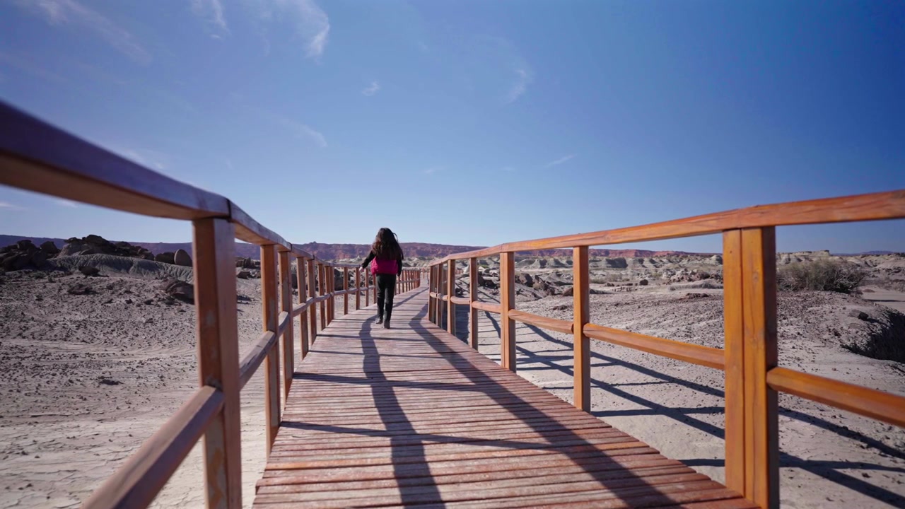 Una donna con uno zaino rosso cammina lungo un ponte di legno illuminato dal sole nel paesaggio roccioso e arido del Parco Provinciale di Ischigualasto sotto un cielo azzurro e limpido