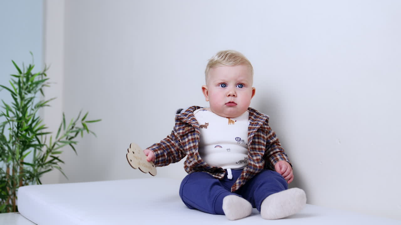 Lovely plump blond baby boy in checkered jacket sits indoors. Cute infant plays with wooden sign. White backdrop.