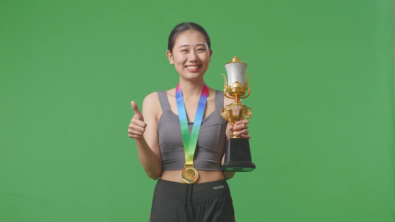 mujer asiática con una medalla de oro y un trofeo mostrando el pulgar hacia arriba y sonriendo a la cámara como la primera ganadora en el fondo de la pantalla verde en el estudio