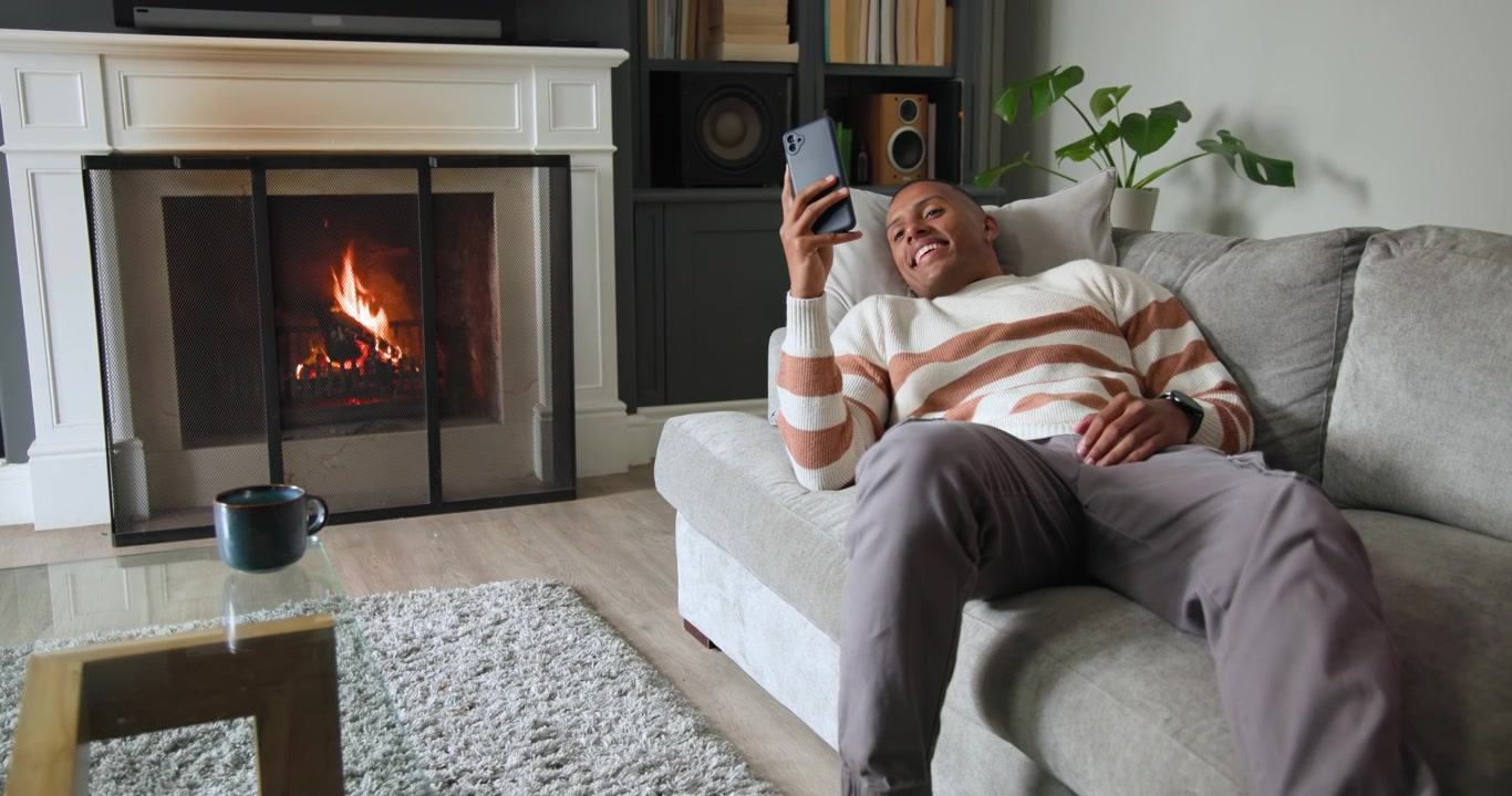Uomo afroamericano in maglione che si rilassa su un divano a destra, alzando il telefono per controllare i messaggi accanto al caminetto.