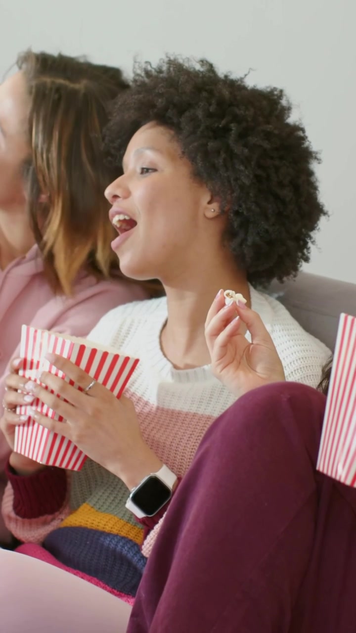 Video verticale: Donna al centro che mangia popcorn da secchielli sul divano, indossando un maglione, davanti allo schermo.