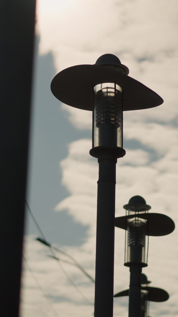 Primer plano de farolas contra un cielo nublado, siluetas de faroles antiguos en postes altos, cables aéreos que atraviesan la suave luz del amanecer, poca profundidad de campo y ambiente minimalista tranquilo, paneo cinematográfico lento