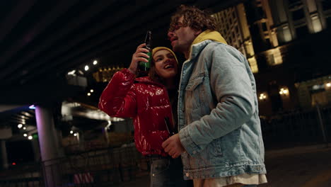 joyful couple laughing under bridge. woman pouring beer into man mouth in city.