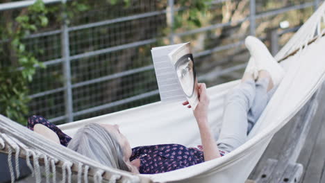 Mujer-Caucásica-De-Mediana-Edad-Leyendo-Un-Libro-Relajándose-En-Una-Hamaca-En-La-Terraza-En-La-Naturaleza,-Cámara-Lenta