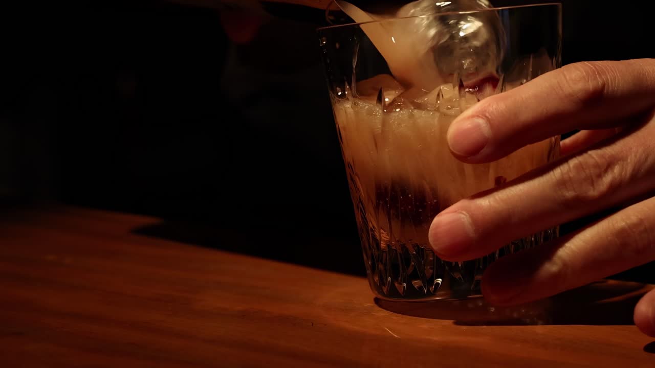 Um bartender habilmente serve uma bebida cremosa e adiciona uma cereja de guarnição num ambiente com pouca luz.