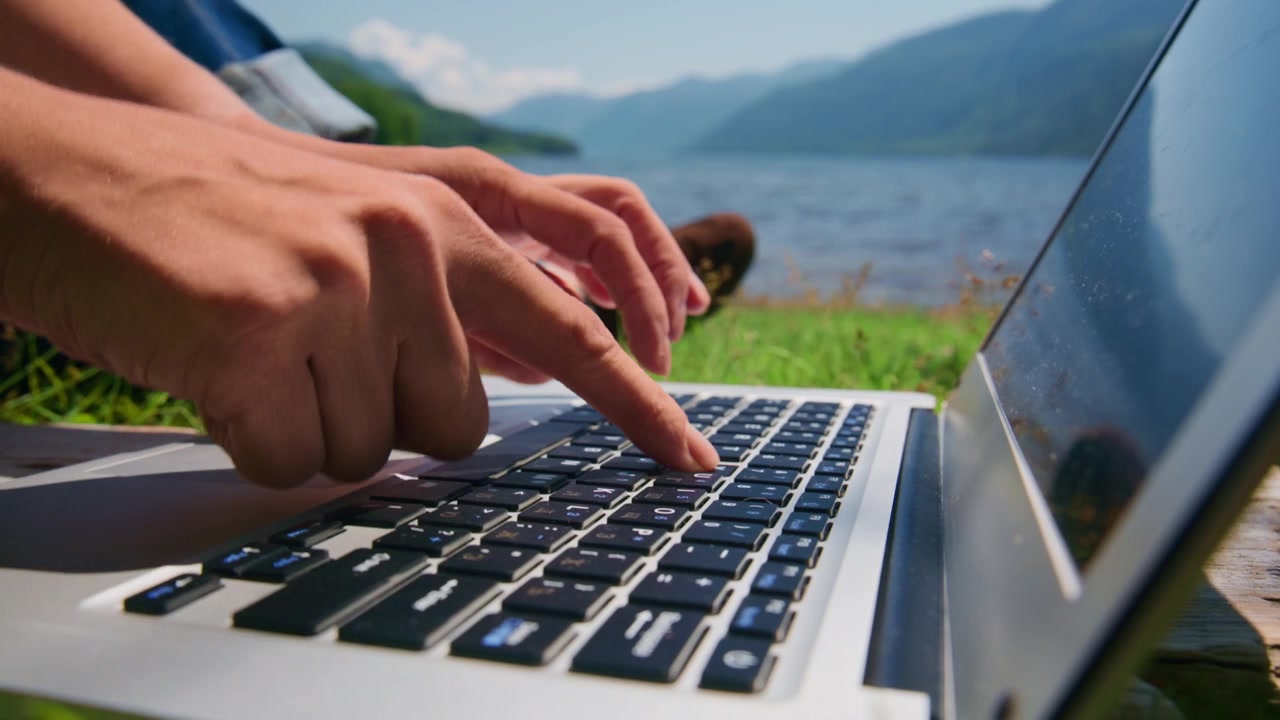 Hands type on laptop keyboard, outdoors near lake. Mountains and green grass behind. Person focused, calm, working remotely. Perfect for remote work, travel, or nature-themed content