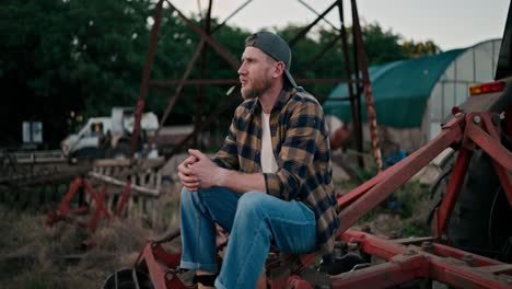 Side-view-of-a-confident-guy-farmer-in-a-cap-holding-a-spikelet-in-his-teeth-and-sitting-on-a-combine-among-greenhouses-on-a-field-on-a-farm