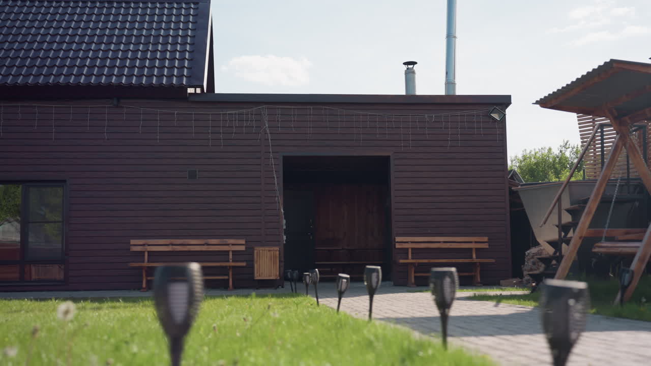 Brown wooden house in sunny fenced compound with paved walkway, bench seating, decorative hanging lights, glass windows reflecting parked car and adjacent cabin, lush grass in foreground