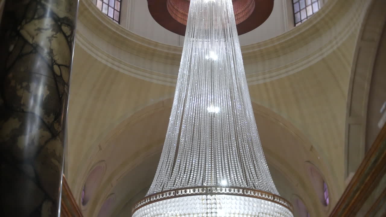 Un grande lampadario di cristallo pende da un elaborato soffitto ad arco in un interno lussuoso, proiettando una luce calda su colonne di marmo—ideale per hotel di lusso, eventi di alto livello o pubblicità di architettura storica