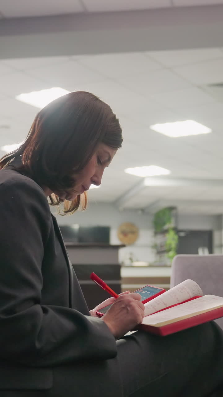 Businesswoman reading notebook in office lounge area. Female professional focused on work or study while sitting on sofa with modern office design in background. Calm and quiet workspace atmosphere