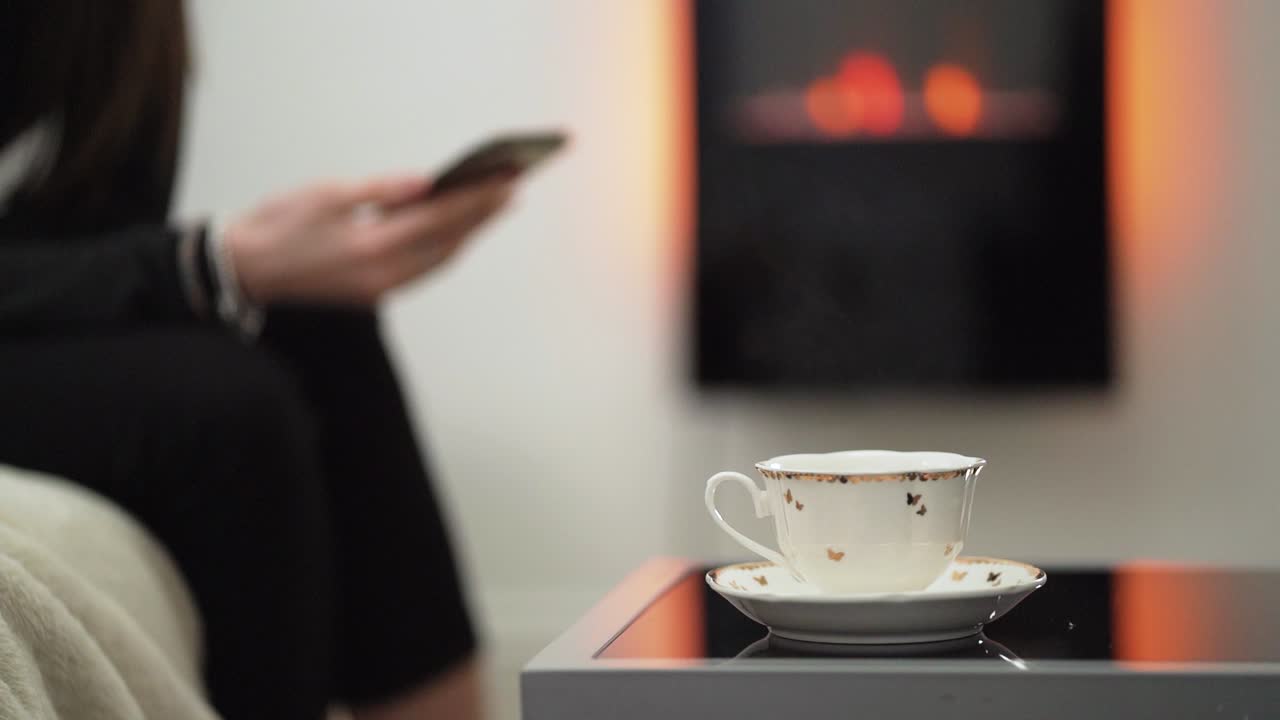 여자가 백그라운드에서 휴대 전화를 사용하는 동안 김이 나는 찻잔 | 클로즈업 샷