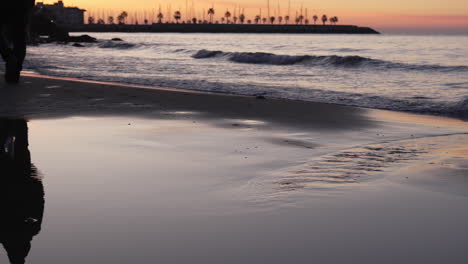 sunrise skyline is reflected in coastal waters that wash up onshore as a man walks across the beach line during golden hour