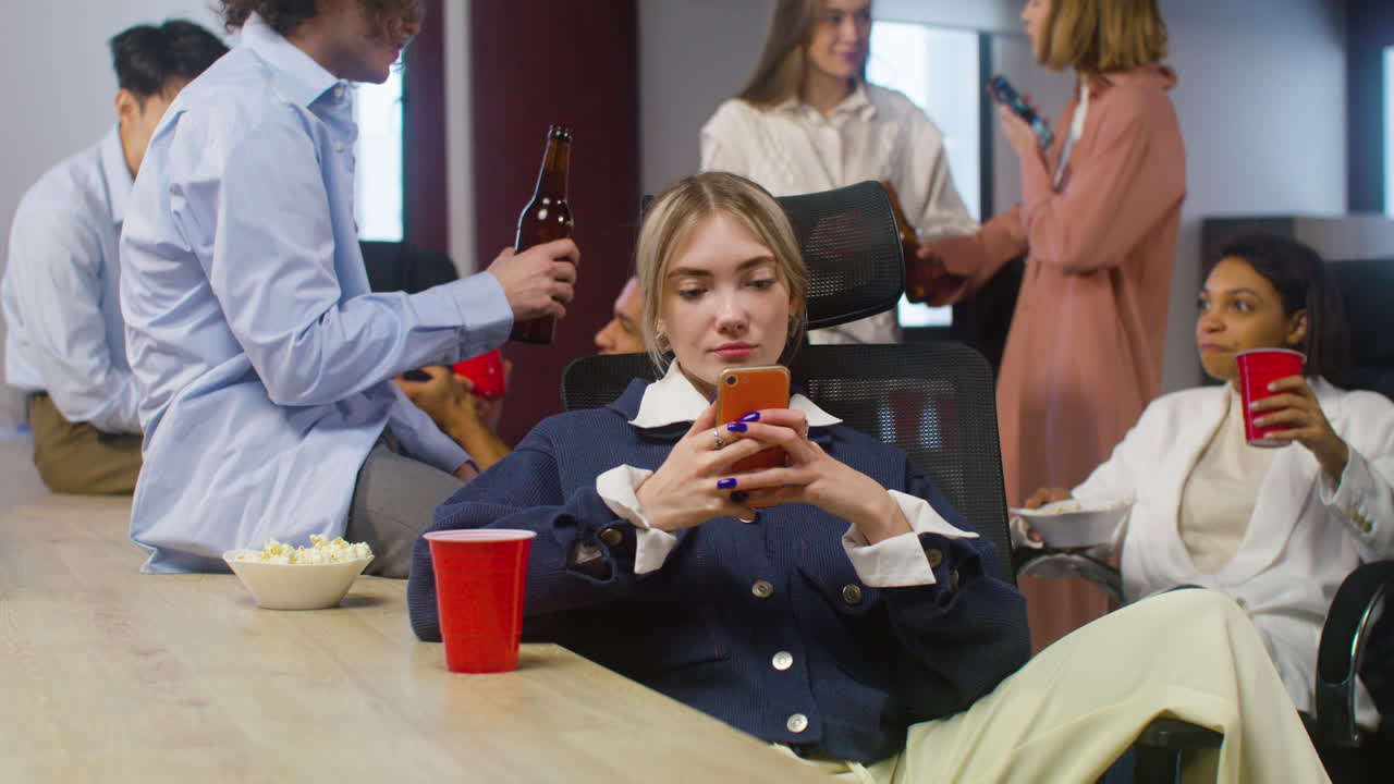блондинка сидит и пользуется мобильным телефоном, в то время как ее коллеги разговаривают и поднимают тосты за выпивку на вечеринке в офисе