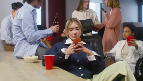 chica rubia sentada y usando el teléfono móvil mientras sus colegas hablan y brindan bebidas en la fiesta de la oficina