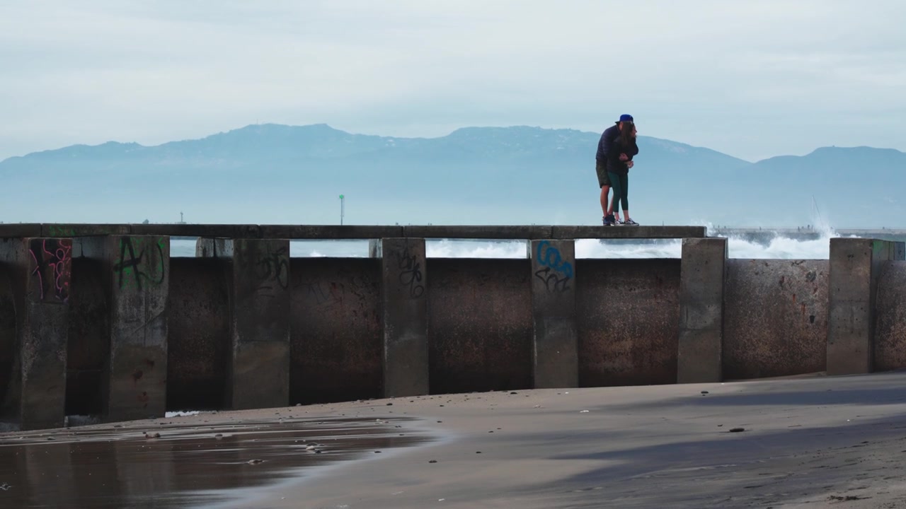 Lykkelig forelsket par klemmer på brygge foran røffe havbølger på Playa del Rey, California, USA