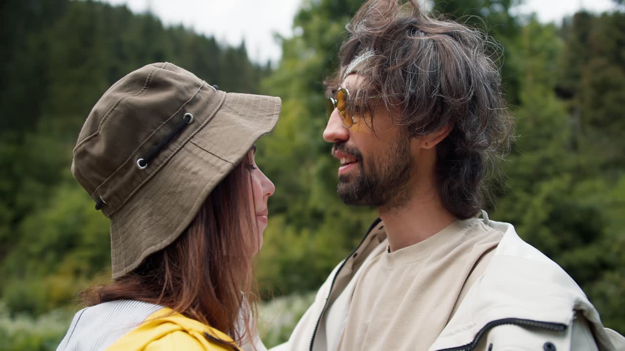 Shooting up close: a guy and a girl are standing next to each other, looking at and straightening each other's clothes. Happy couple in hiking clothes on green forest background