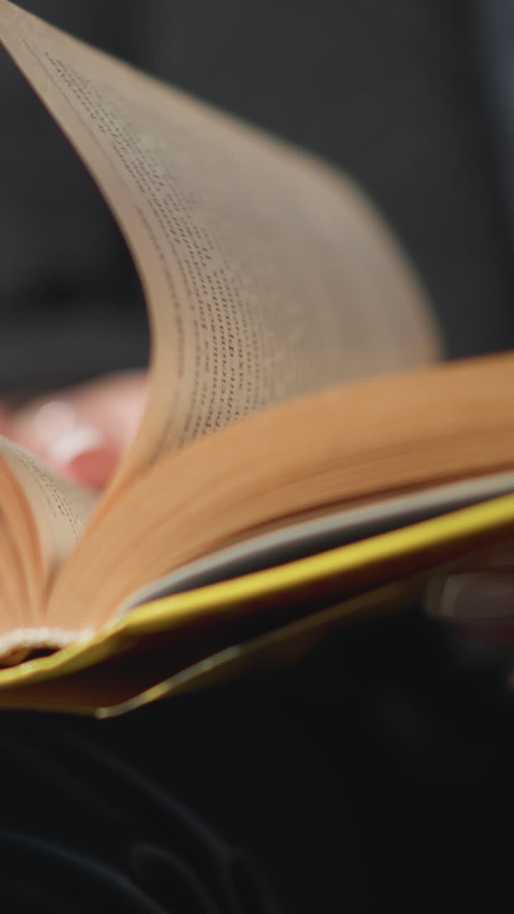 Close-up of book pages being flipped by a hand adorned with a ring and polished nails, dressed in black clothing, the background is blurred