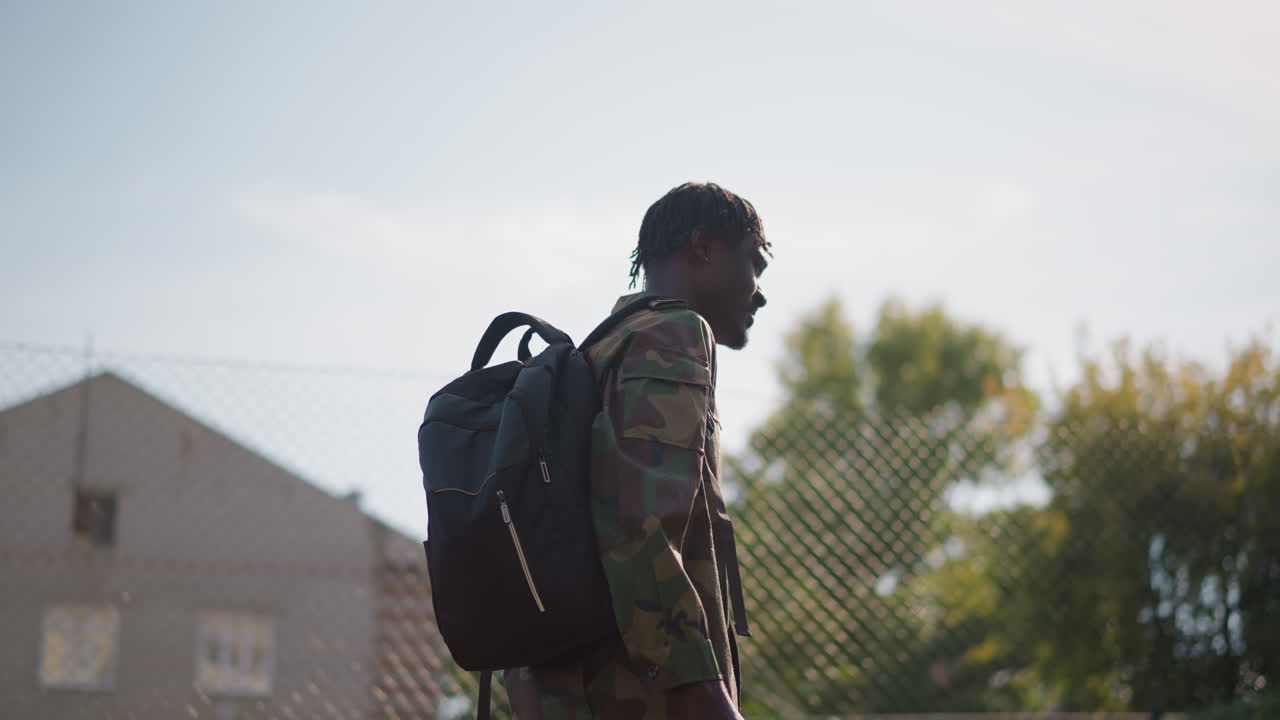 Black Soldier Walking With Backpack Fence Backdrop, Urban Suburb Under Warm Sunlight. Camouflage Uniform And Contemplative Posture Convey Homecoming Mood And Quiet Resilience. Wide And Close Shots