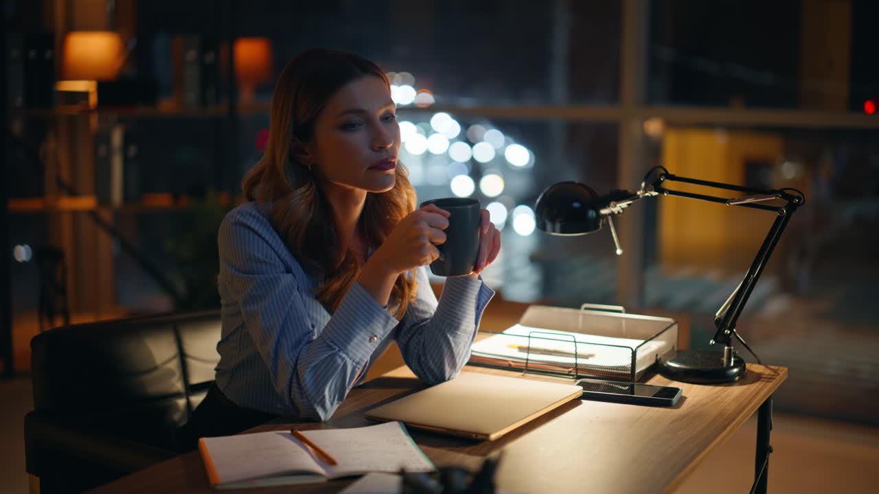 Manager assonnato che beve caffè, stanco per il lavoro straordinario, seduto sul posto di lavoro sotto la luce della lampada.