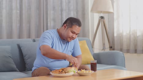 a fat asian man dipping french fries in ketchup eating fast food on a sofa in the living room at home