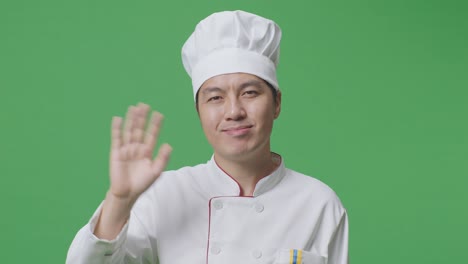 close up of asian man chef smiling, waving hand, and saying bye while standing in the green screen background studio