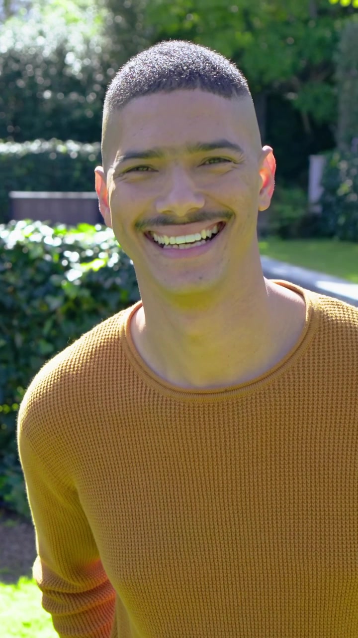 Video verticale: Uomo sorridente con maglione a maglia color senape che si gira verso la telecamera e ride in un cortile soleggiato.