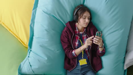 estudiante feliz usando un teléfono inteligente navegando en línea mensajes de redes sociales disfrutando de la pausa para almorzar relajándose en almohadas coloridas escuchando música en una oficina moderna vista superior