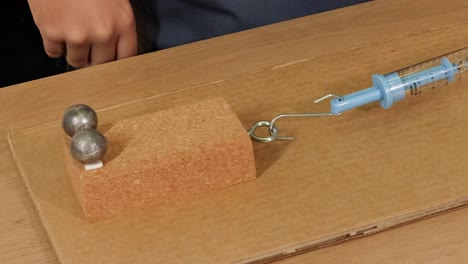A cork block with metal weights is pulled across a cardboard surface using a blue spring scale.