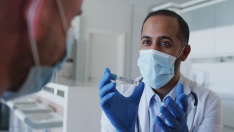 Mixed-race-male-doctor-wearing-face-mask-consulting-male-patient-at-home-before-vaccine