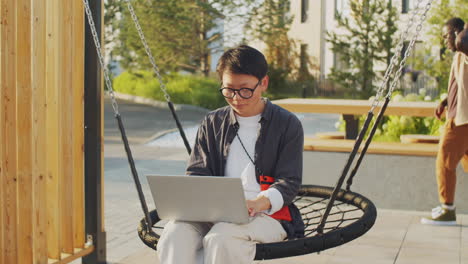 asian woman sitting on swing outdoors and working on laptop