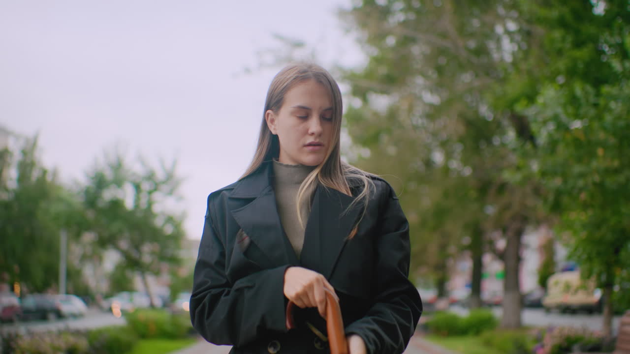 Female hidden behind large black umbrella as she close it standing on city street with blurred background of trees cars and buildings during cloudy day showing outdoor lifestyle