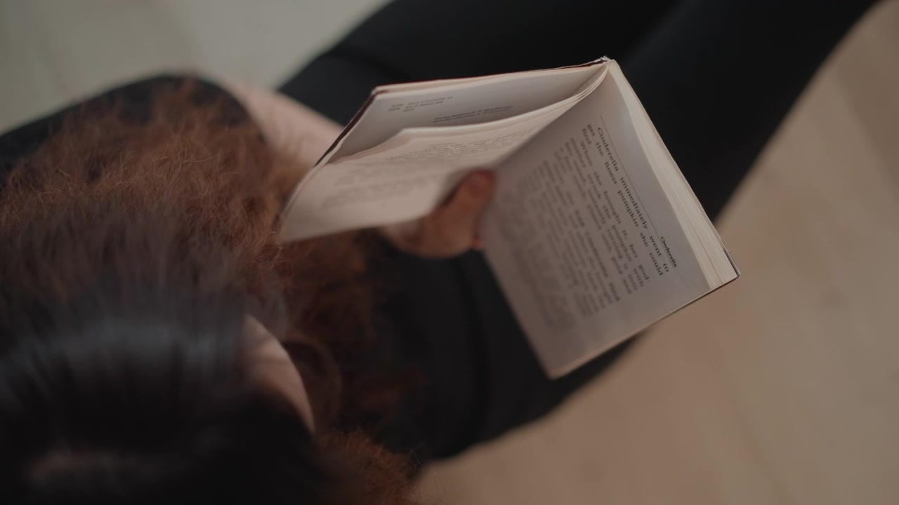 Scena dall'alto di una persona seduta che tiene un libro aperto, con la sommità dei capelli castano scuro al centro e una leggera sfocatura della mano che suggerisce un delicato voltare pagina, in una luce interna calda, con lettura, libro, dall'alto, capelli.