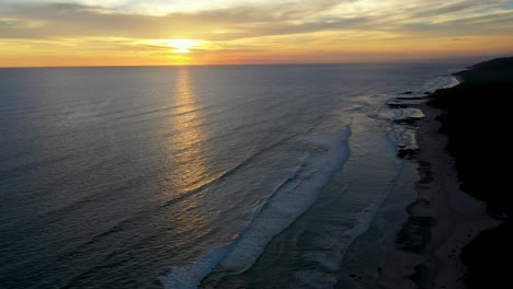 Antena-De-Hermosa-Puesta-De-Sol-Reflejándose-En-El-Agua-Con-Olas-Y-Surfistas-En-El-Océano-Pacífico-En-Tamarindo,-Costa-Rica