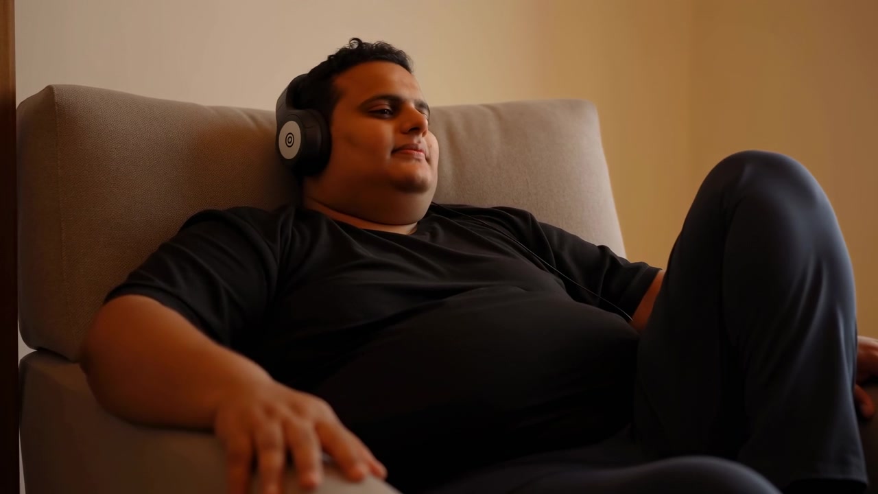 Adult male reclines in beige fabric armchair wearing wired over-ear headphones, eyes closed and slight smile, creating calm listening mood inside warm interior with soft light and neutral decor