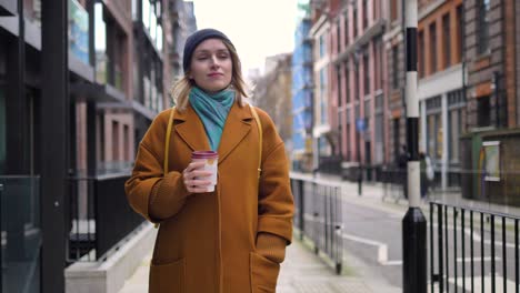 feliz alegre positiva y relajada joven mujer caucásica disfrutando del tiempo, caminando por una calle de londres con una taza de café en las manos, y sonriendo mientras mira a su alrededor