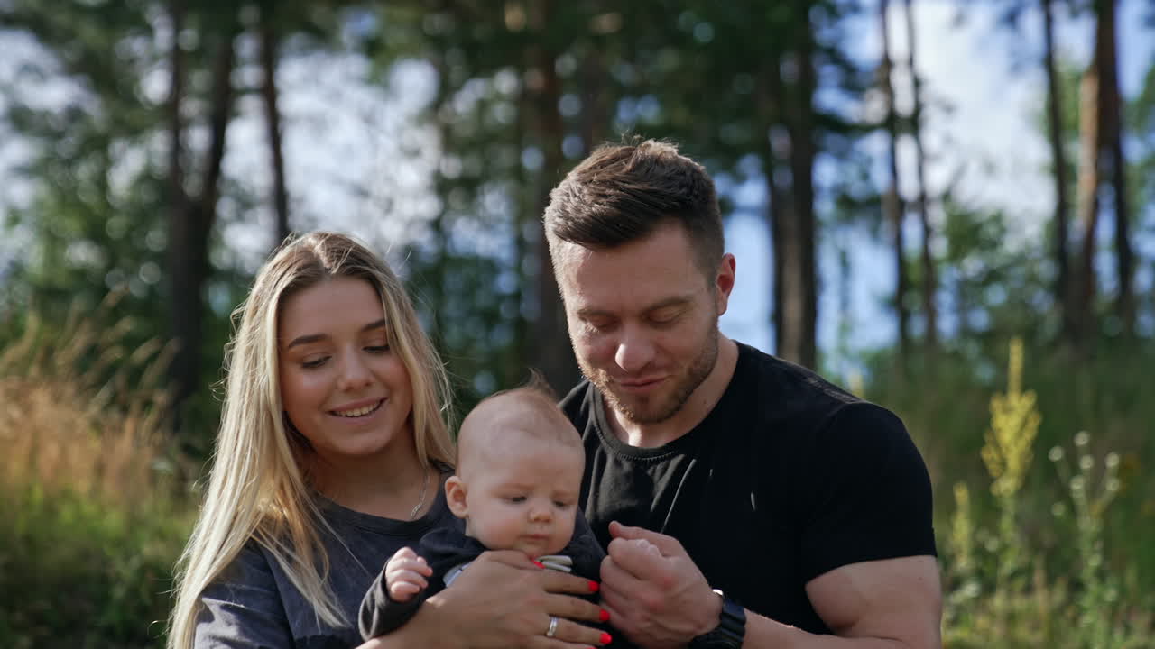 Adorable blonde newborn is held by his loving parents. Happy family having leisure time in nature on sunny summer day.