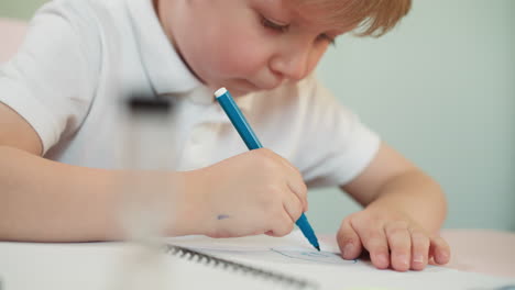 Little boy puts pen cap and draws picture in copybook at table