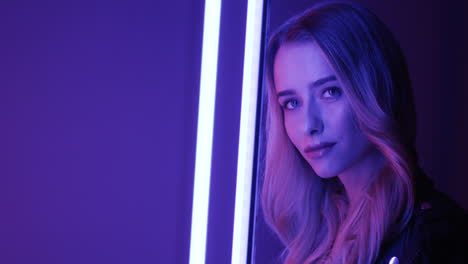 close up view of blonde young woman standing among neon lights and smiling cheerfully to the camera