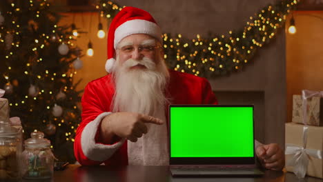 In-the-background-of-the-Christmas-tree-an-elderly-Santa-Claus-is-holding-a-laptop-and-pointing-at-the-screen-with-a-chromakey.-Laptop-with-a-green-screen.-Santa-looks-at-the-camera.-High-quality-4k-footage