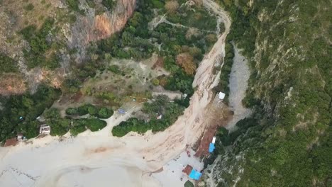 Hermosa-Agua-De-Color-Y-Olas-Del-Mar-Adriático-Chocando-Contra-La-Playa-De-Gjipe-Con-El-Impresionante-Cañón-De-Gjipe-En-El-Fondo-Y-Las-Enormes-Montañas-De-Albania-Al-Atardecer