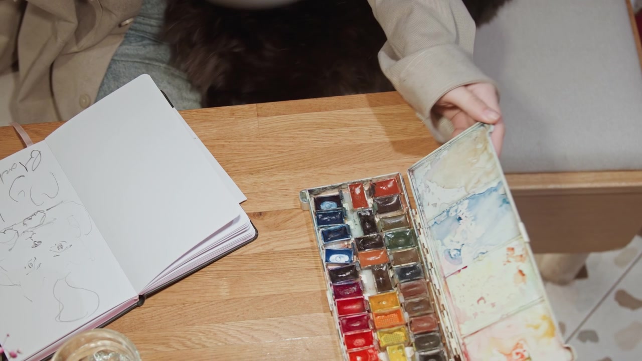 Primo piano delle mani di una persona che aprono colori ad acquerello su un tavolo di legno da artista con un album da disegno, un bicchiere e un vaso nelle vicinanze. Un gatto nero giace sullo sfondo per tutta la scena. Una luce naturale soffusa illumina.