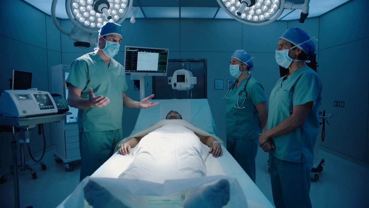 Clinico capo in camice che fa un briefing, gesticolando verso il paziente in sala operatoria e preparandosi per l'intervento chirurgico con i monitor.