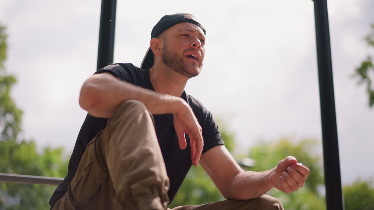 White Man Sitting Outdoors Checking Phone, Casual Skater Taking Break On Steps, Cap Backwards, Pensive Gaze, Sunlit Park Background, Handheld Smartphone, Candid Microexpressions, Waiting For Friend