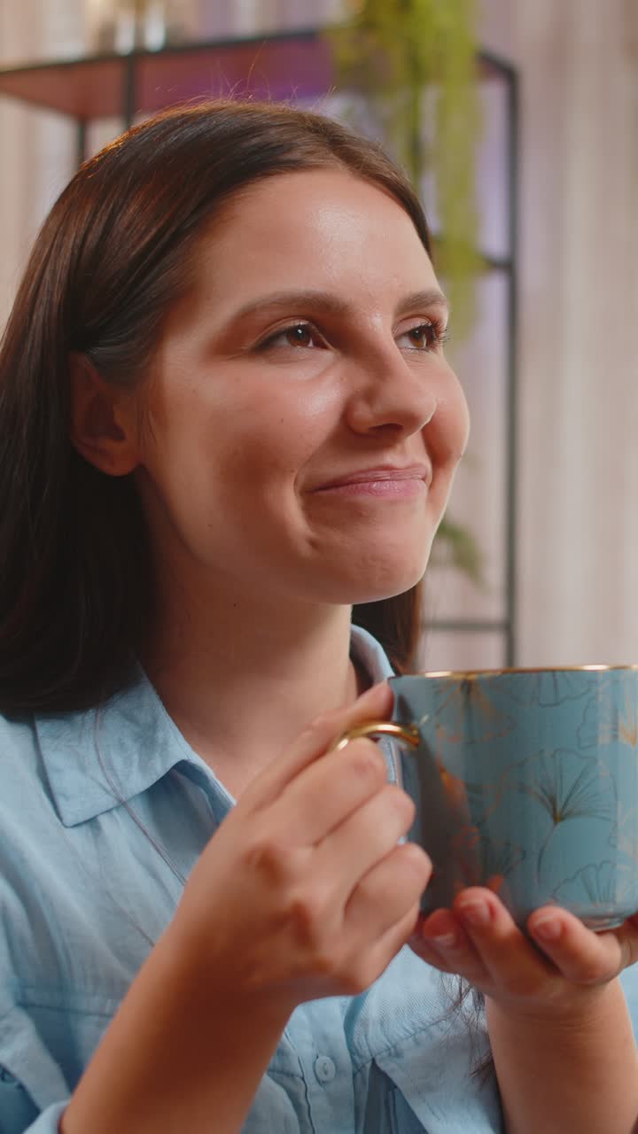 Ragazza giovane attraente e sorridente che beve una tazza di caffè o tè alle erbe seduta sul divano di casa