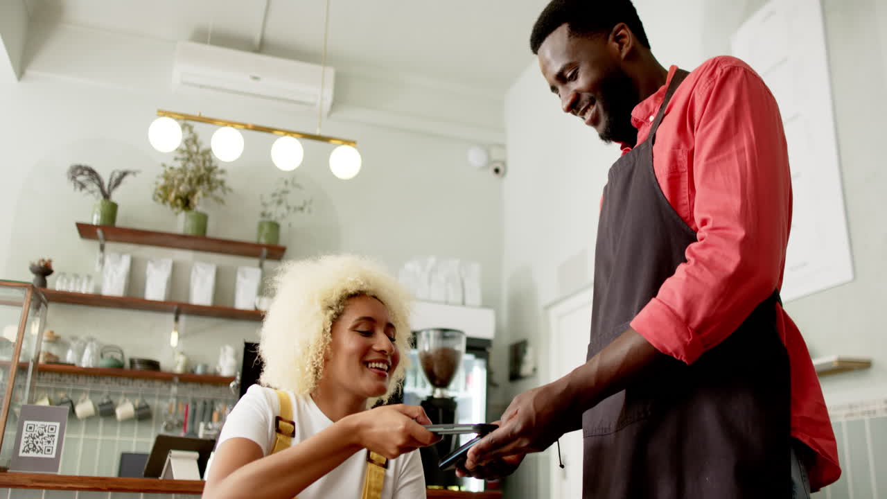 A young biracial woman is paying with a smartphone to an African American worker in a cafe