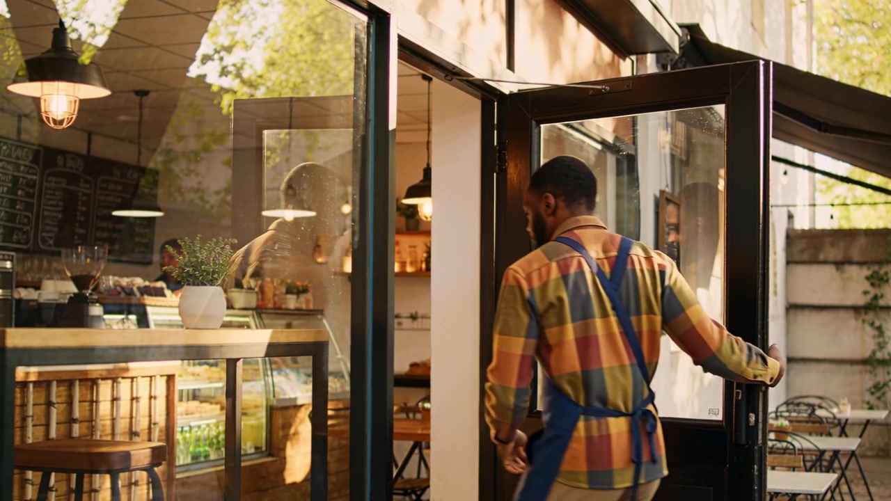 Uomo con grembiule blu che si avvicina e apre la porta a vetri per entrare in un caffè di quartiere, con spazio per il testo.