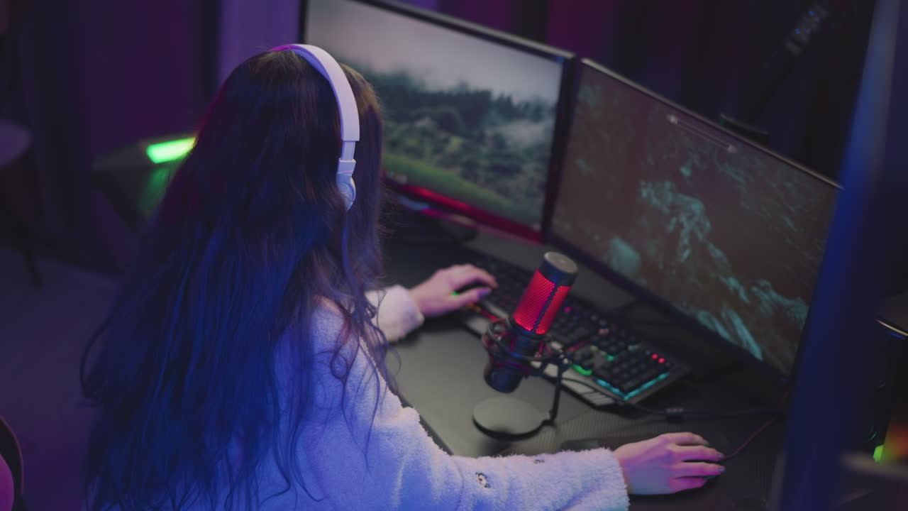young woman with long wavy hair wearing white headset and fluffy sweater intensely playing game on dual monitors with red microphone and illuminated keyboard in dark colorful gaming room