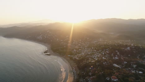 Amplia-Antena-De-Amanecer-Con-Destellos-De-Sol-Sobre-Una-Pequeña-Ciudad-De-Surf-Costera-Mexicana,-4k