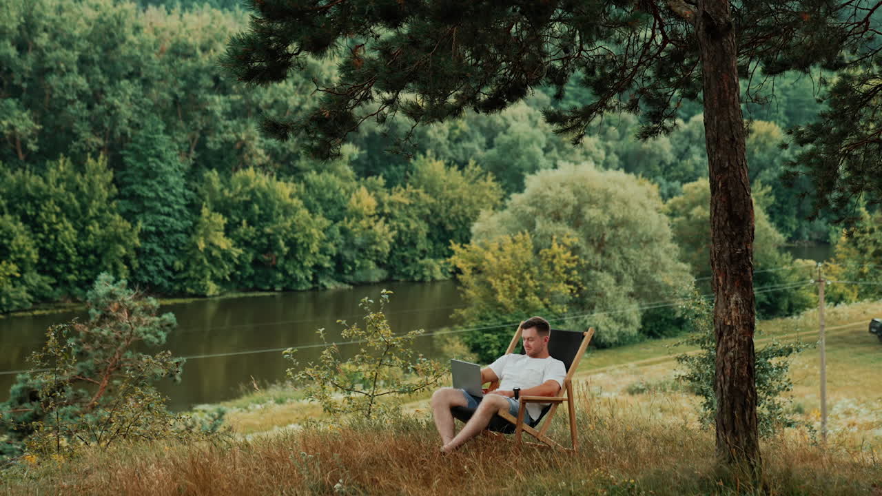 Man sits in a folding chair on the hill over the river. Freelancer works remotely on his laptop in the nature.